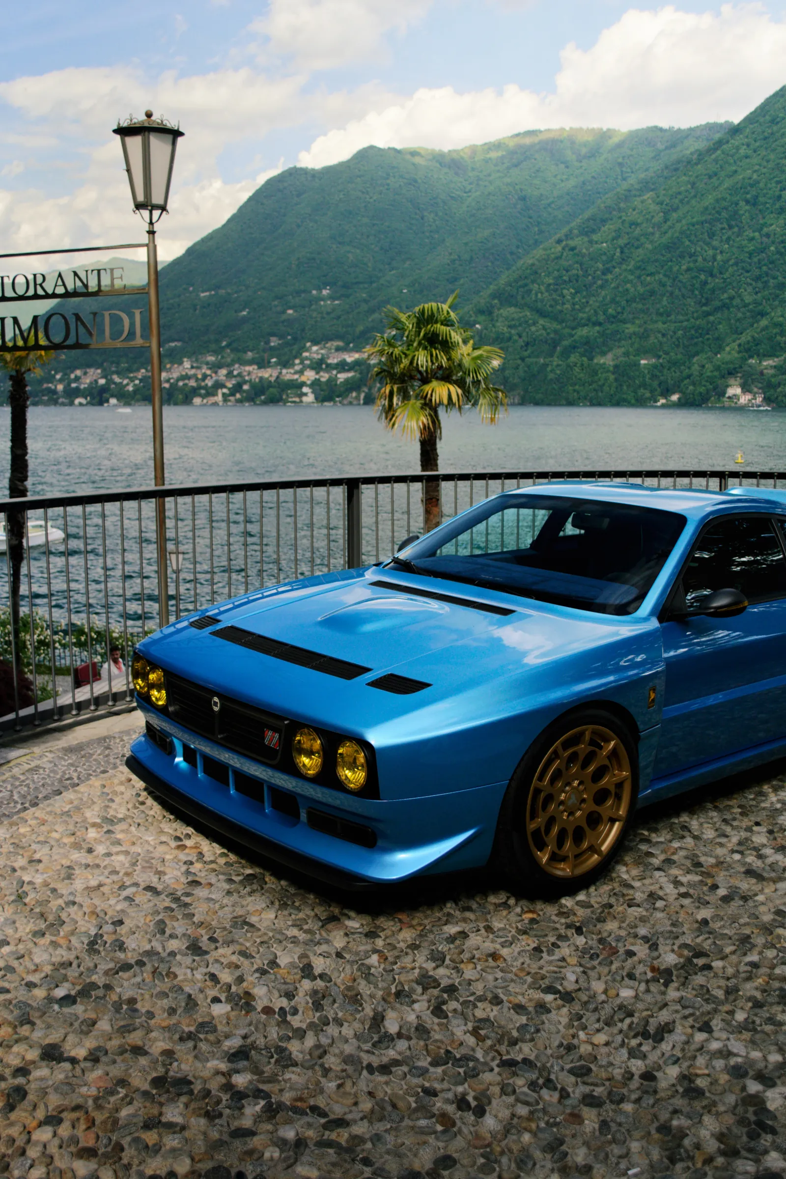 Auto sportiva retrò blu con cerchi dorati accanto a un lago con montagne